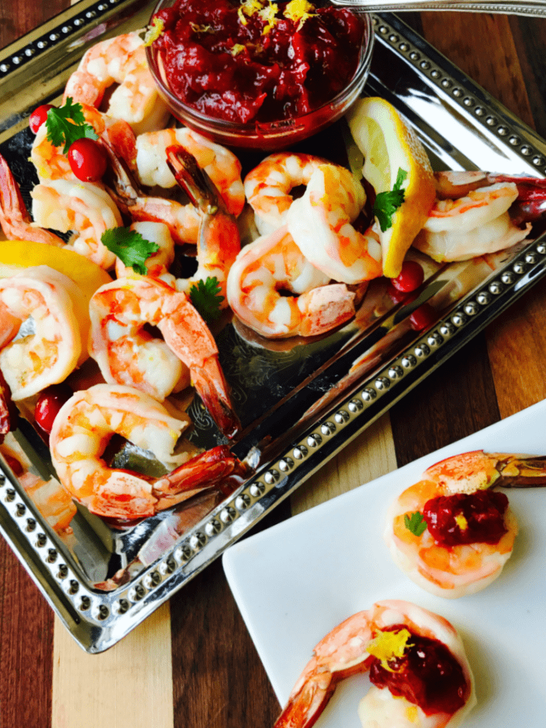 Cranberry Horseradish Chutney in a glass bowl with shrimp cocktail on a platter. On the side is a white plate with two shrimp topped with chutney and lemon zest.